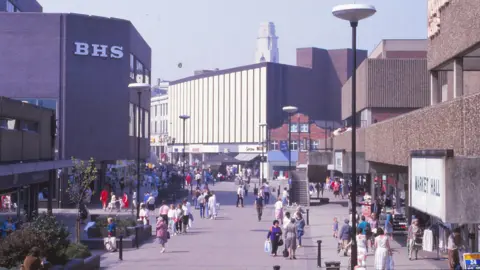 Barnsley Archives Barnsley's busy pedestrian shopping street lined with large concrete buildings in a modernist style. On the left, there is a building with a large BHS sign, and on the right, a sign reads Market Hall. The walkway is wide and paved, with many people walking and shopping. Tall round street lamps line the street, and in the background, a white building with vertical black stripes and a clock tower are visible.