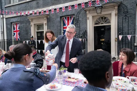 PA Media Keir Starmer apprehensively pours a teapot into a teacup held by a person wearing military uniform, with other attendees around the table that contains various bits of afternoon tea in front of Number 10 Downing Street. Union flag bunting adorns the building.