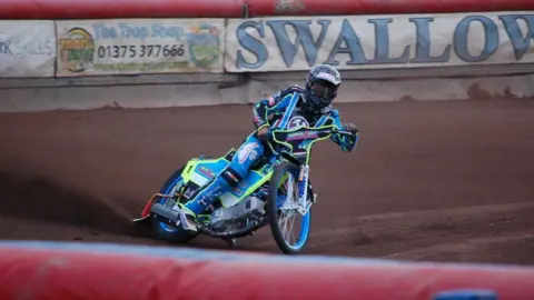 A speedway rider in action on a blue and neon yellow motorbike. The helmet and protective gear they are wearing matches their bike and also contains various logos from advertising companies. The rider looks to be skidding to a halt with dirt spraying to the left of the image from the back wheel of the bike. 