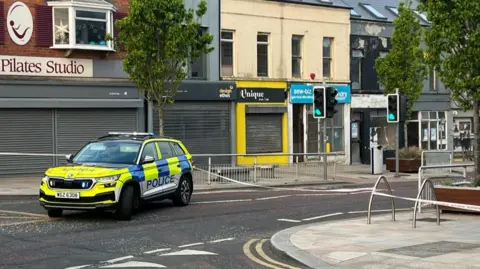 A yellow and blue PSNI vehicle is parked in the middle of the road. A police cordon lines the top of a high street. A number of shops in the background have their shutters down.