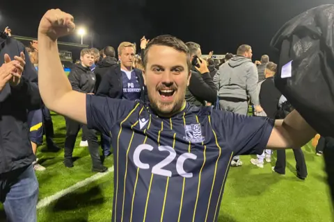 Warren Muggleton/BBC A man wearing a dark blue Southend United jersey is raising his fists cheering in front of the camera. Behind him are other young men crowding on the pitch at Forest Green Rovers.