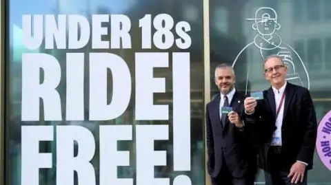 South Yorkshire Mayor Oliver Coppard and councillor Sir Steve Houghton CBE hold a Barnsley Micard. Two men stand side-by-side in dark suits, holding small rectangular cards. They stand in front of a large window with the word printed 'Under 18s Ride Free!'.