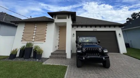 Chynna Perkins A house with windows temporarily hidden by Kevlar coverings, and with a vehicle parked outside 