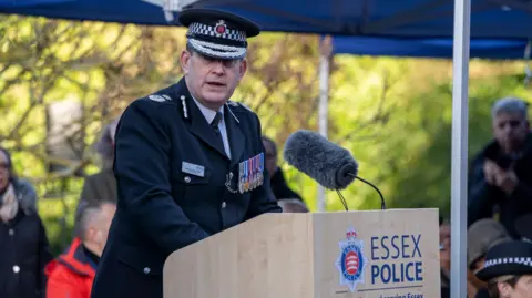 Essex Police Ben-Julian Harrington wearing a black policing uniform and hat with a line of medals pinned to his chest. He is speaking behind a lectern which has a large microphone on it. People watch on behind him.