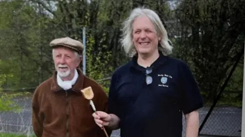 Boz Davison Man in a brown jacket with white facial hair and a flat cap, standing with a man in a ark t-shirt with long white hair holding a spell (wooden stick) to revive the traditional northern game of Knurr and Spell for Yorkshire Day