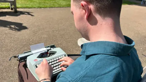 A man in a blue shirt seen from behind as he balances a typewriter on his knee, typing words on a small piece of paper on a sunny day. He is sitting on a park bench and another park bench can be seen beyond, with grass and trees behind it.