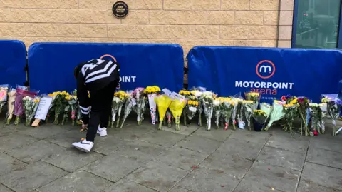 A woman next to dozens of flowers laid beneath a circular black plaque in Adam Johnson's memory