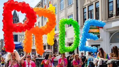 Mark Kerrison/Getty Images A drag queen and four ladies in pink tops walk down the street holding a different letter of the word pride and each in a different colour