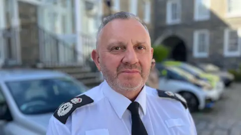 BBC Damian Kitchen - A man with short grey hair and a stubbly grey beard, wearing a white shirt and a black tie with Police insignias on his shoulders on black pieces of fabric. It is a police uniform, without the jacket. 
