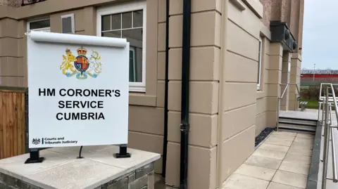 The entrance to the coroners court in Workington. There is a sign outside indicating this is the coroner's court. There is a ramp with metal rails leading to the main door. The building is painted beige.