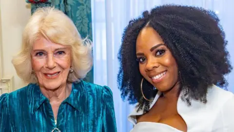 Selina Brown, on the right, has curly black hair to her shoulders and wears a white top while smiling. Queen Camilla, on the left, has shorter blonde hair and smiles, wearing a turquoise dress.