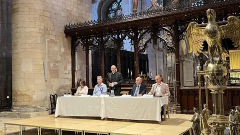 bbc Four people sitting either side of a standing vicar on a stage in Tewskesbury Abbey next to a large golden eagle lectern