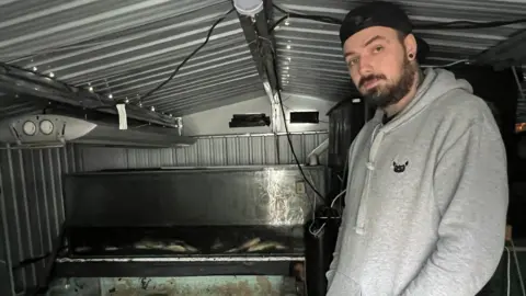 LDRS Craig Anderson, who is wearing a grey hoodie and black snapback cap, stands inside a shed next a counter of dead fish above a tank.