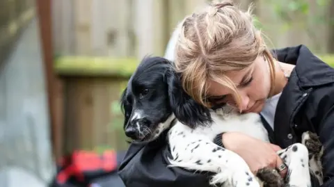 Black and white coloured Puffin being held in the arms of his owner Tabitha. She is resting her head on the dog's shoulder. She has blonde hair which has been tied back and she is wearing a black coat.