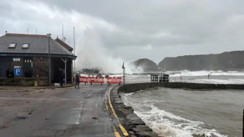 SKatieShore A wave crashes over old stone harbour. It is cordoned off with red and white barriers. The sky is grey and the water is murky. 