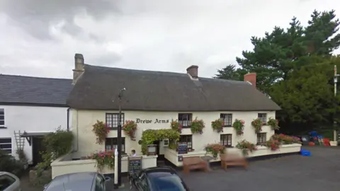 Google Exterior of The Drewe Arms. The cream pub has a thatched roof. It has eight windows across the front of the building. A red sign on the building states Drewe Arms. A number of benches are in front of the pub. 