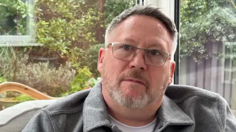 A man with greying hair and a beard, who wears glasses and a grey shirt, sits in front of a window looking onto a garden behind.