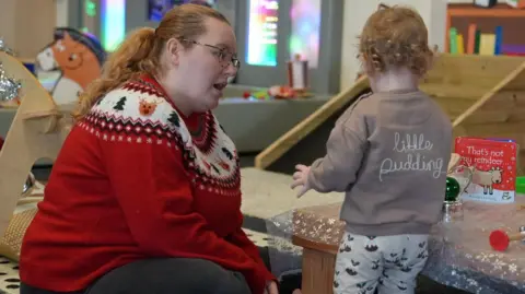 BBC Lucy Jackson-Hind with her one-year old son at the Clart About Sensory Cafe in Consett. Ms Jackson-Hind is sitting on the carpet, side-on to the camera and is talking to the toddler, pictured from behind, standing next to a low table with some toys and books. Ms Jackson-Hind has strawberry blond hair tied back in a ponytail. She is wearing a red Christmas jumper with reindeers and Christmas trees. Her son has short blond, curly hair and is wearing a grey jumper with the words little pudding on the back and grey trousers with images of Christmas puddings.