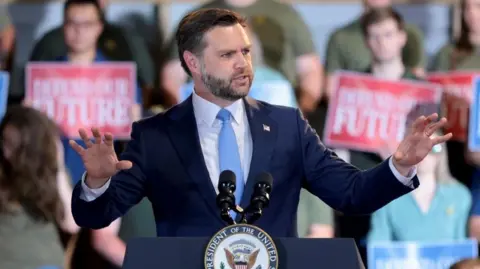 Getty Images Vice-President JD Vance addresses a crowd of supporters in Maine. He is wearing a blue suit and holding his arms out, with a number of supporters sat behind him holding signs saying "Defend our Future"