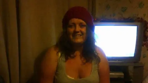 Family photo Tanya smiling to camera with shoulder length dark hair curled at the ends. She's wearing a maroon beany hat and a grey vest top. The photograph is in a living room setting with a TV behind her left shoulder.    