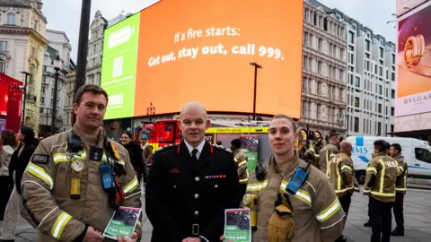 PA Media London Fire Commissioner Jonathan Smith stands in front of large billboards with two colleagues - one male and one female - either side of him