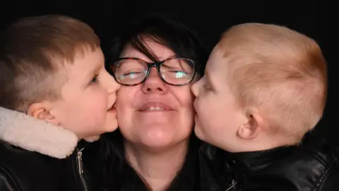 Handout for the family A dark-haired woman with glasses smiles at the camera while her two young sons, both with short blonde hair, give her a kiss on the cheek.