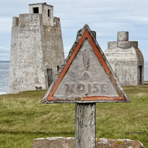 Fiona Finlayson A faded triangular sign warning about noise, in front of disused foghorn structures.