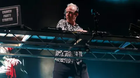 The image shows a DJ performing on stage at a live music event. The DJ is standing behind a large metal-framed table with professional DJ equipment, including mixers and turntables. There is a laptop or controller on the left side with a visible sticker or label that reads “CRAVEN PARTY STARTER.” The DJ is wearing a patterned black-and-white top and dark trousers. Overhead stage lighting creates blue and white spotlights shining down and some haze or smoke effects in the background.