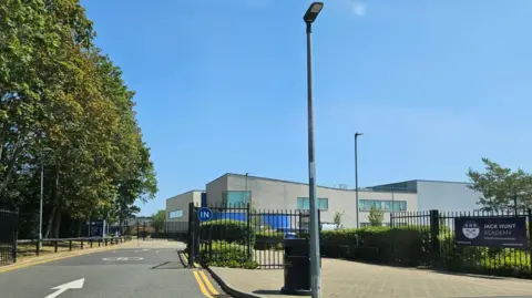 Shariqua Ahmed/BBC An image of a school, which is a big white building with "Jack Hunt School" written on it in silver, and black gates in front of a car park.