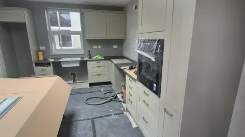 An olive green kitchen with black worktops and large gold handles which is half-renovated and built with protective polystyrene and cardboard around some parts of it. There is a window at the back above the sink.