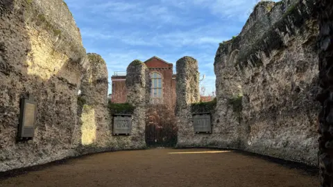 Three stone walls sit around an open courtyard. There is a gate in the wall at the back. The sky is clear and bright blue.