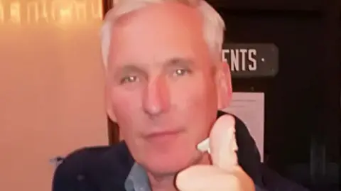 Family handout Jim Paterson is giving a thumbs up to the camera while standing outside a toilet door. He has short, white hair and is wearing a grey shirt and dark jacket.