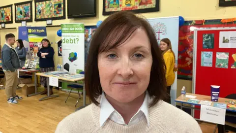 A woman wearing a cream coloured sweater with brown hair looking at the camera. Behind her are information stalls set up in a school hall 