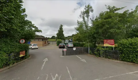 Google Google Street View of Lytchett Matravers Primary School gates. The wide vehicle entrance is lined with trees and shrubs. Three cars can be seen on the tarmac driveway beyond the gates.