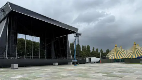 A large pop up stage outside. It is in the process of being assembled. To the right is a large blue and yellow tent. In front of the stage is a grey flooring system. There is a blue crane completing work on the stage. 