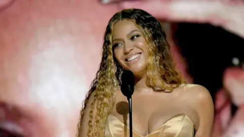 Beyonce accepting an award and speaking into the microphone. She's wearing a gold strapless dress and smiling 