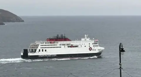 The Manxman ferry, which is painted in the Isle of Man Steam Packet Compnay colours of white, red and black and has STEAM-PACKET.COM written in the side. It is sailing across Douglas Bay on an overcast day.