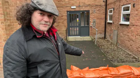 BBC Chris Cooper pointing at sandbags outside his flat on Waterside Close, in Loughborough, Leicestershire.