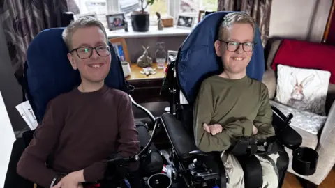 Identical twins Sam and Alex Bolton pictured sat in their wheelchairs in their living room smiling. 
