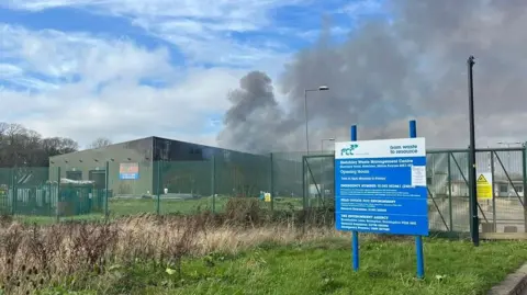 Holly Nichols/BBC A close up of a fire at a recycling centre. A large plume of grey smoke rises from the top of the building, with bits of corrugated metal showing signs of damage.