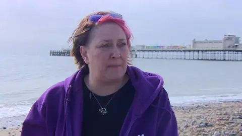 A woman with a pink streak in her hair looks sad on a beach. 