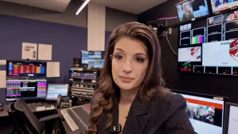 A woman sits in a video control room wearing glasses and looking into the camera.