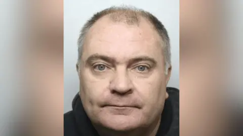 A mugshot of Green, who has short, thinning hair on the top of his head, with more hair visible on the sides. The hair appears to be a mix of grey and dark tones. He is wearing a dark-coloured hoodie, which is visible around the neck and shoulders. The background is plain and light-coloured.