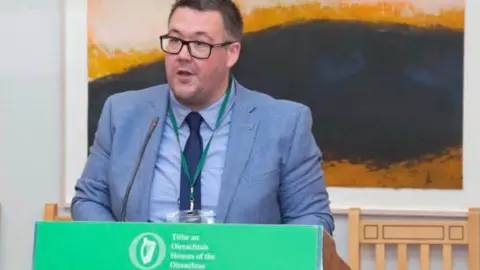 Jason Poole Jason Poole. He has short dark hair, wearing a light blue blazer, light blue shirt and navy tie. He is standing at a podium with Houses of the Oireachtas branding on it. A piece of artwork is behind him.
