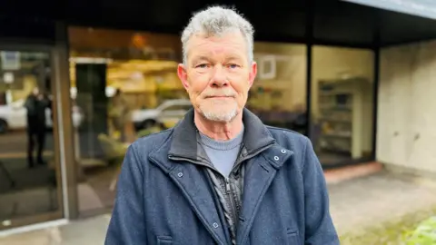 Man with grey hair and grey stubble looking at camera, wearing a blue jacket and jumper, standing in front of a library building.