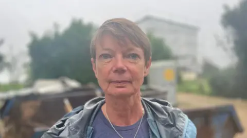 Sarah Horton is a woman with short brown hair. She has blue eyes and is staring into the camera. She is wearing a blue t-shirt, a grey waterproof jacket and a silver chain necklace. Behind her the background is an out-of-focus skip, some trees and a house covered in scaffolding.