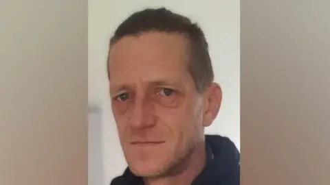 Head and shoulders shot of Vadims Zmicerevskis who has short hair and is wearing a black zipped up jacket and coat. There is a beige wall in the background.