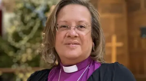 Patricia Hillas, who has shoulder-length light brown hair and is wearing rimless glasses. She dressed in a purple shirt with a clerical collar and black clerical smock.