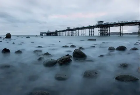 Llandudno pier at sunrise, taken from the sea. Image shows the rising tide with black rocks and the pier in the distance with a pink sky behind