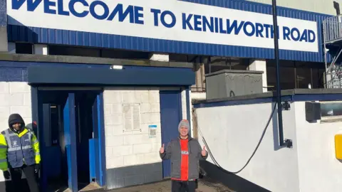 Mark Crowther A man wearing a Luton shirt and a woolly hat is stood in front of Kenilworth Road with his thumbs up